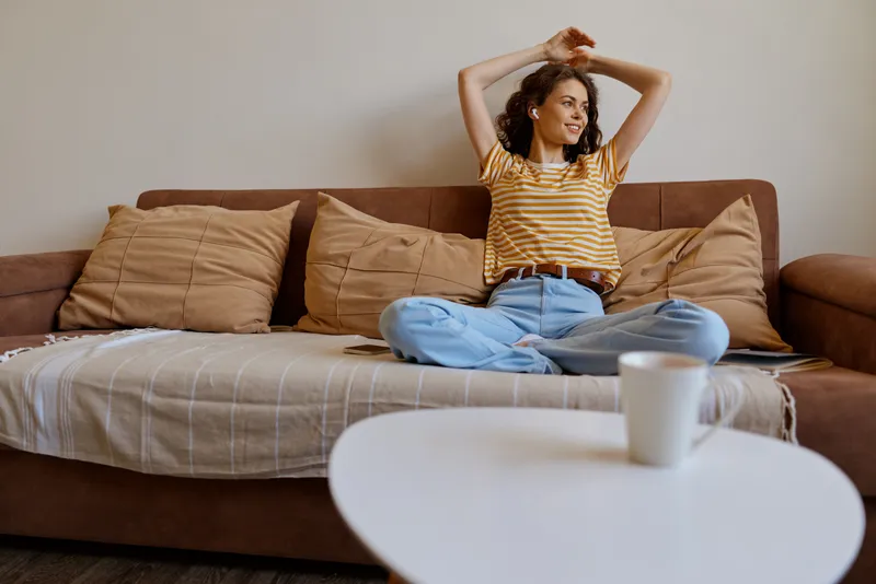 A young woman sits cross legged on a brown sofa, stretching her arms overhead with a relaxed content smile, casually dressed in a striped top for a cozy home lifestyle scene