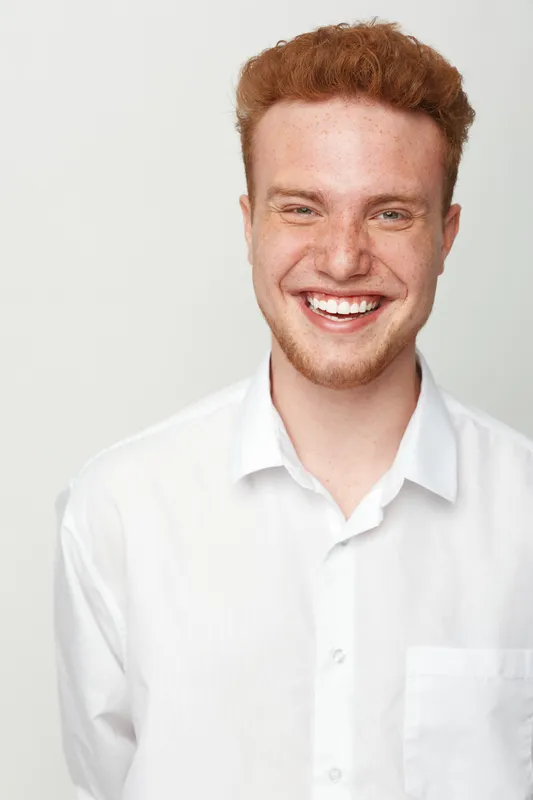Smiling young man with red hair and beard wearing white shirt stands against white background. Portrait of happy male model with natural expression and casual style for lifestyle concept.