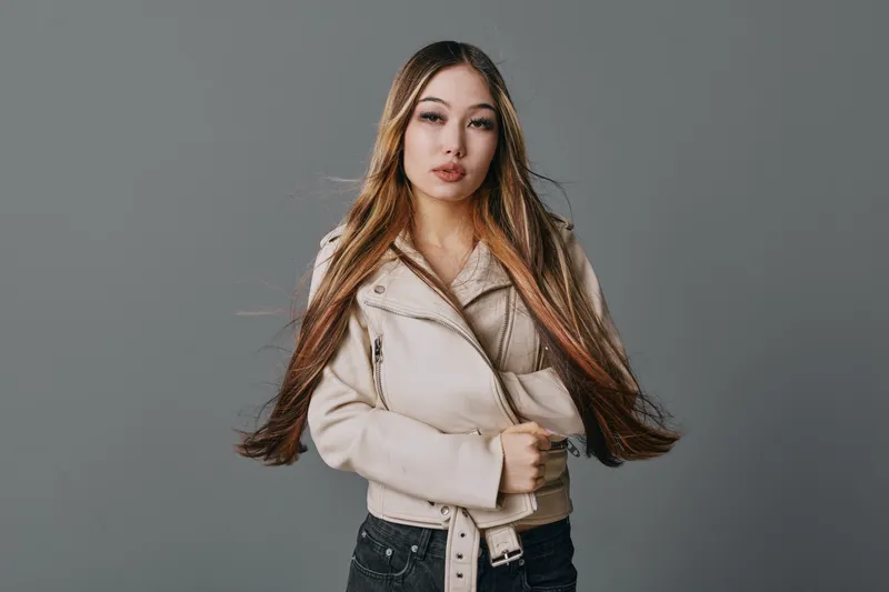 Young woman with long straight hair wearing beige jacket standing against grey background looking at camera with confident expression and relaxed posture in studio portrait.
