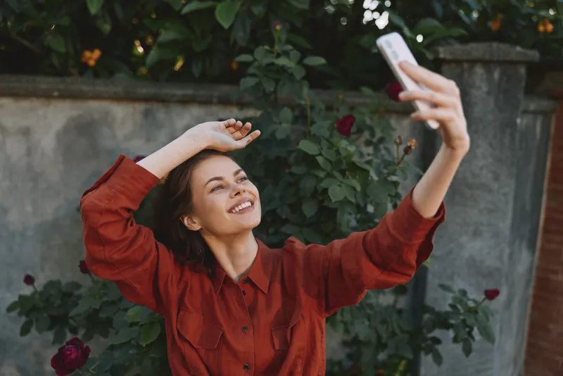 A joyful woman in a red shirt takes a selfie outdoors, smiling with arms raised in a garden backdrop. A candid lifestyle moment of confidence and happiness