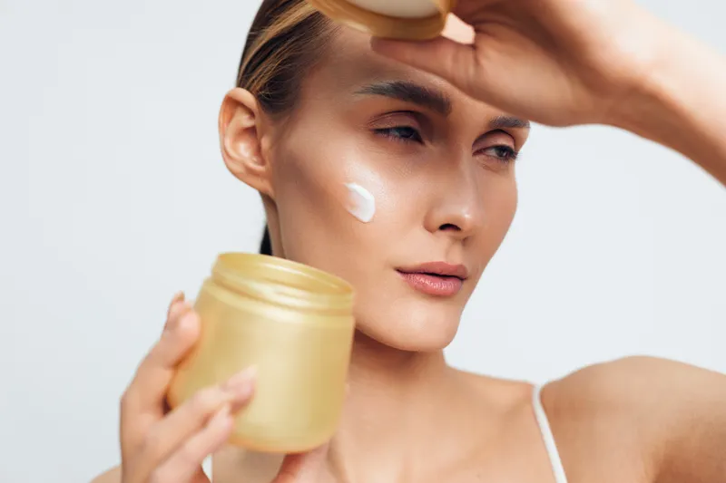 Young woman with clear skin applying white face cream on cheek holding a gold jar against a light background. Concept of skincare, beauty, wellness, and daily self-care routine.