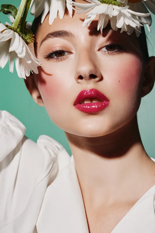Beautiful woman with glossy red lips and delicate daisy flowers on her head. Close-up fashion portrait on green background. Concept for beauty, fashion, and creative makeup.