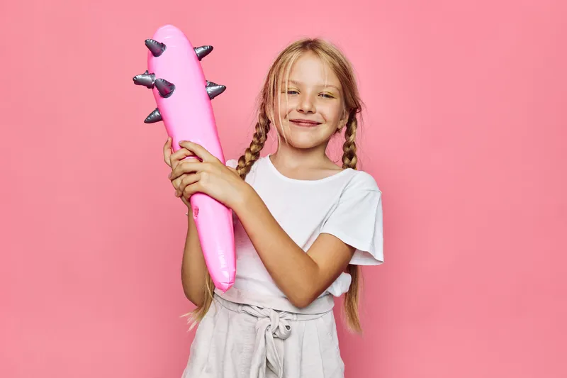 Happy girl with long hair in white shirt holding inflatable pink toy with spikes against pink background. She smiles confidently while posing in studio lighting.
