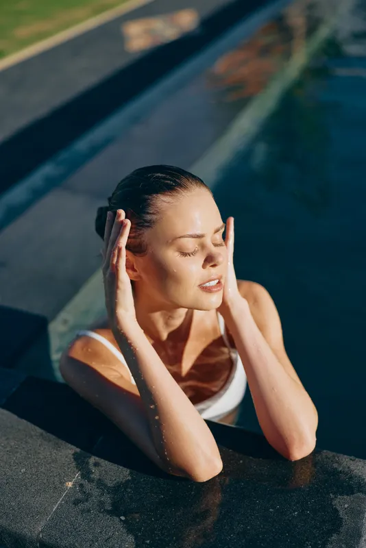 Young woman relaxes in outdoor pool with eyes closed, enjoys sunlight and warm water, has peaceful expression, lifestyle concept of wellness and leisure.