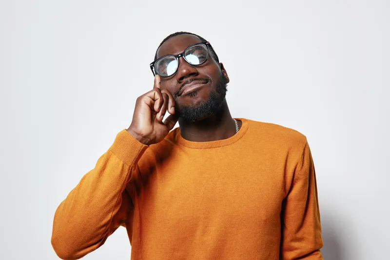 African American man with dark skin wearing glasses and orange sweater smiling with closed eyes and hand near face on white background. Portrait, casual style and positive mood.