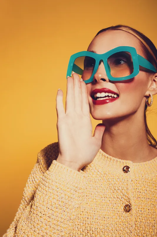 Stylish woman in large turquoise sunglasses and yellow sweater smiles brightly, holding hand near mouth. Vibrant studio portrait with warm yellow background, concept of fashion and joy.