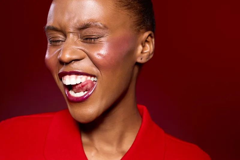 Close-up of joyful African woman with dark skin and vibrant makeup laughing with eyes closed on red background. Concept of happiness, beauty, and expressive emotions in portrait photography.