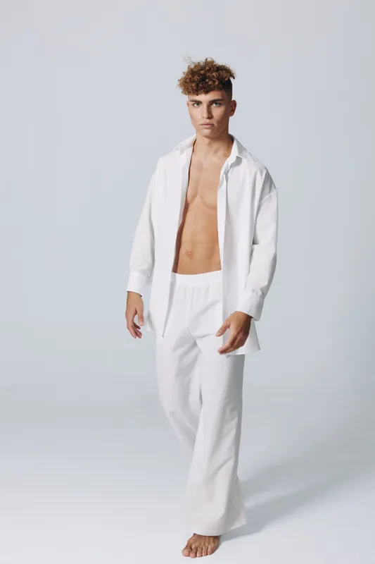 Young man with curly hair wearing white loose pants and open shirt standing barefoot on plain background. Casual fashion, confident pose and minimalistic studio portrait with natural look.