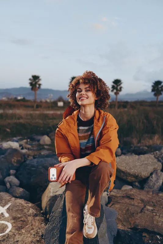 Woman lifestyle outdoors, curly hair, orange jacket, sitting on rocks, holding a smartphone, sunset glow, palm trees, calm smile, travel moment, nature, scenic desert field, dusk