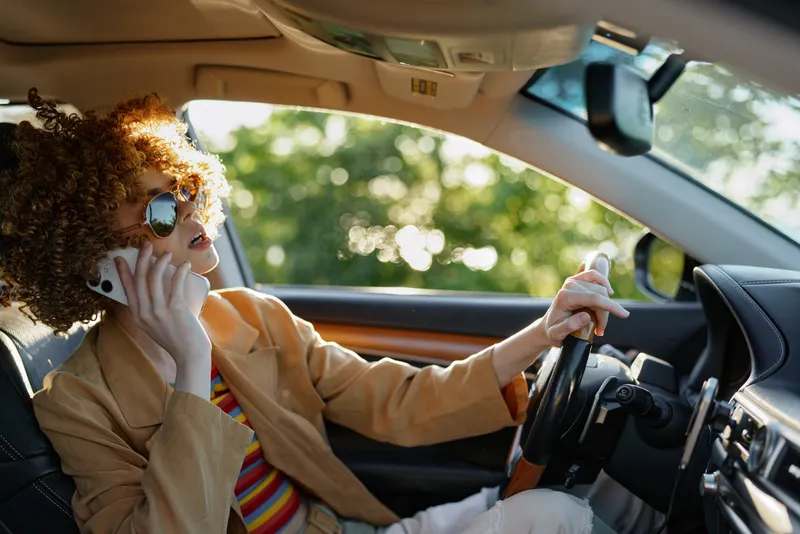 Young woman with curly hair drives a car, talks on a cellphone, wearing retro sunglasses and a striped shirt, bathed in warm nostalgic color.