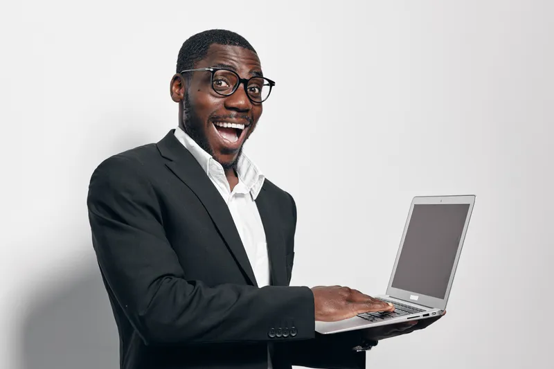 African American man with dark skin wearing glasses and black suit holding laptop smiling and looking excited in studio with white background for technology and business.