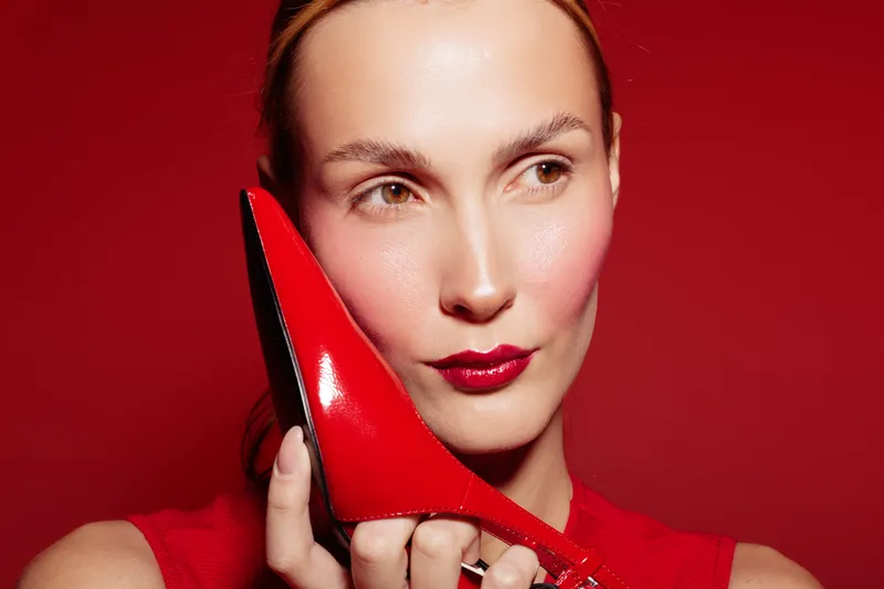 Close-up of a woman holding a red high heel shoe near her face on a red background. Concept of fashion, style, beauty, and elegance with vibrant colors and glossy texture.