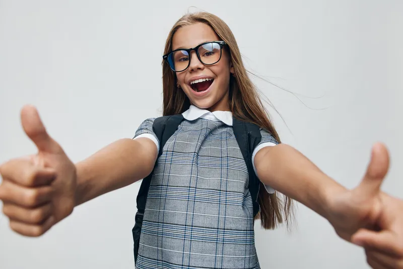 Girl with glasses smiling and giving thumbs up gesture wearing backpack and plaid dress isolated on white background for happy school kid and success.