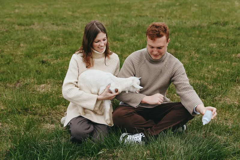 A relaxed couple sits on a grassy meadow, cradling a white goat kid while the man feeds it with a bottle. A warm lifestyle moment showcasing care, bonding, and the simple joys of animal companionship