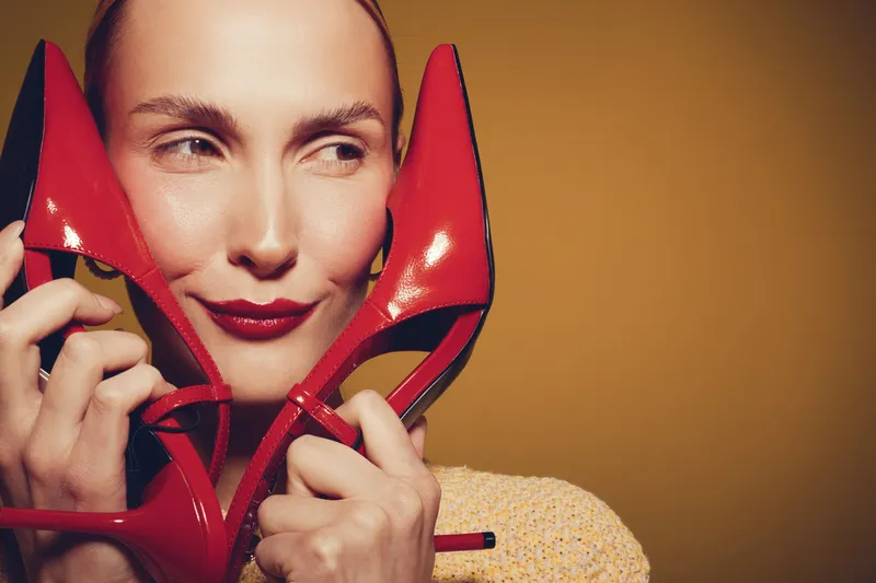 Smiling woman holds red high heel shoes near face, wearing red lipstick and beige sweater. Studio portrait with warm yellow background. Concept of fashion, style, and beauty accessories.