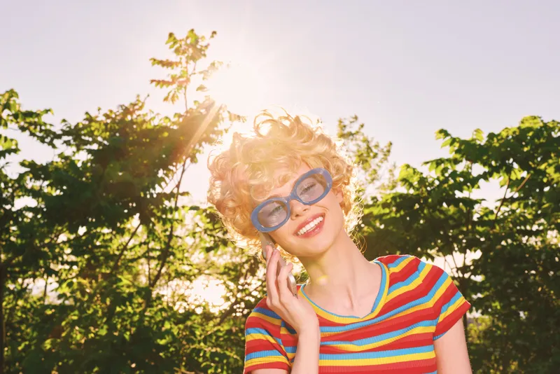 Happy woman with curly blonde hair wearing a colorful striped shirt and blue glasses, outdoors in bright sunlight, retro film color, cheerful lifestyle portrait.