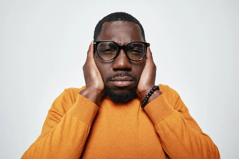 African man with dark skin wearing glasses and orange sweater covering ears with hands, expressing discomfort or stress on white background in studio portrait.