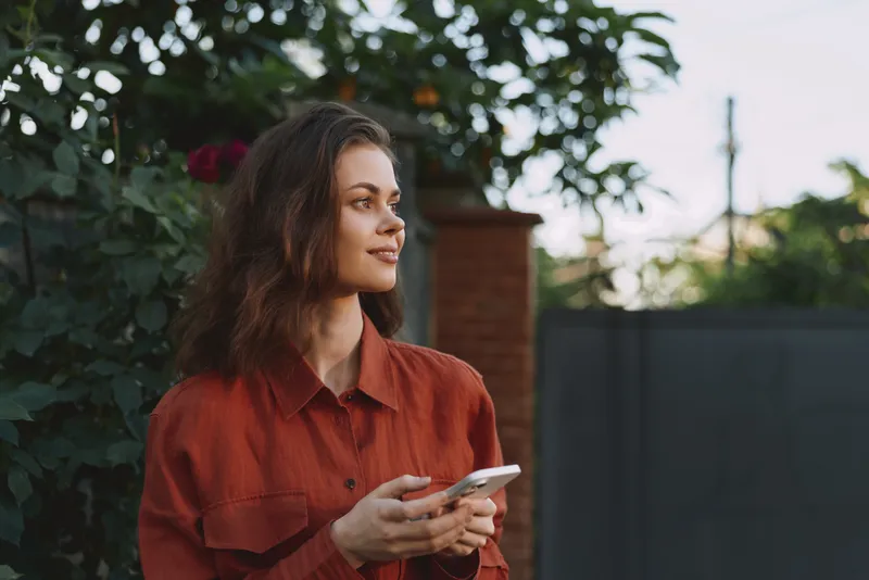 A woman in a rust shirt stands outdoors, smiling softly while holding a smartphone. A casual lifestyle moment captured in natural light and garden surroundings