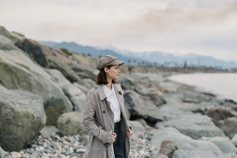 Young woman in warm coat and hat stands on rocky beach looking into distance. Overcast sky and calm sea create peaceful atmosphere, concept for solitude and outdoor lifestyle.
