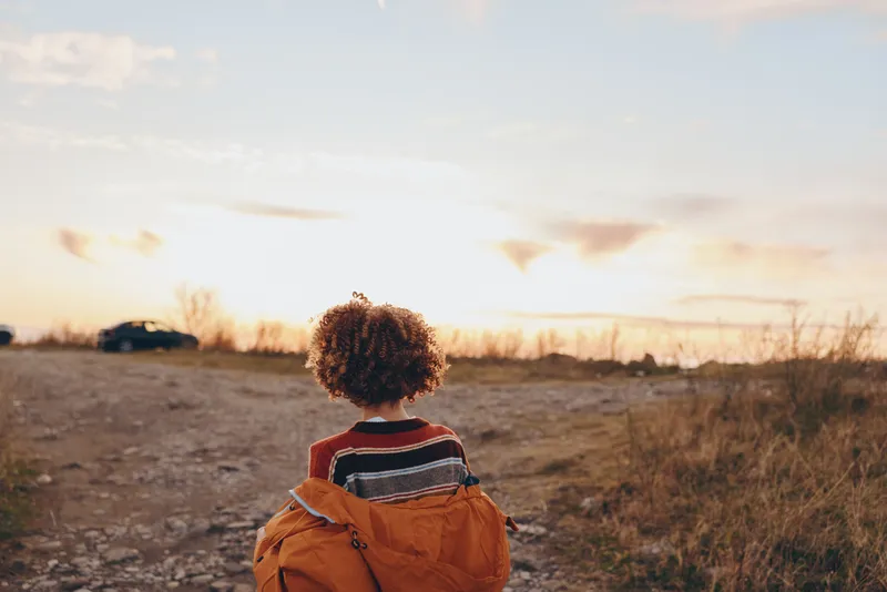 Child outdoors from behind in warm sunset light, wearing orange jacket, facing open rural field, calm lifestyle moment, nature horizon, soft breeze, golden colors, countryside road ahead