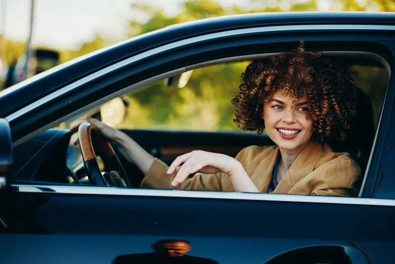 A smiling young woman with curly hair sits behind the wheel in a car, warm vintage tones emphasizing a candid, relaxed urban travel mood.