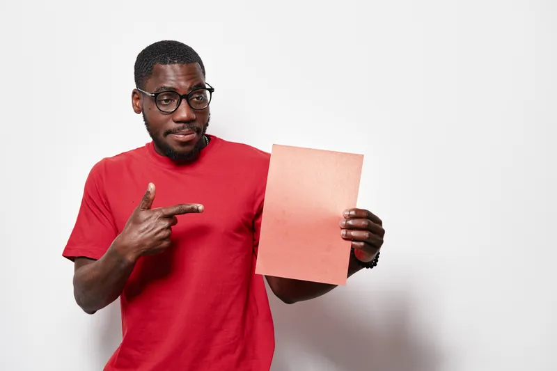 African American man with dark skin wearing red t shirt and glasses holding blank paper and pointing at it on white background for presentation or advertisement.