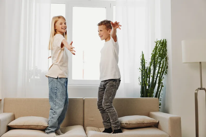 Happy children jumping on couch in bright living room, enjoying playful moment together. Concept of childhood fun, family time, and energetic activity indoors on casual weekend.