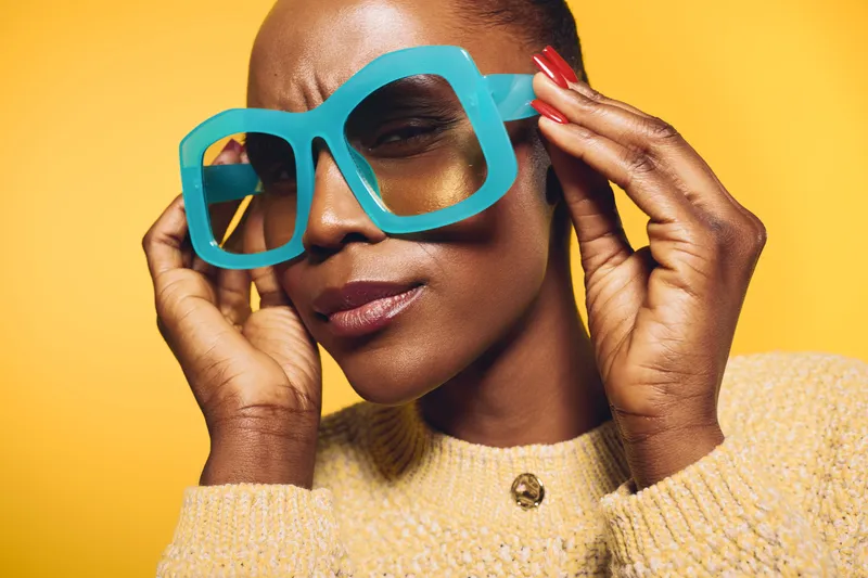Close-up of confident African American woman with dark skin wearing large blue glasses and beige sweater against yellow background. Concept of fashion, style, and beauty portrait.