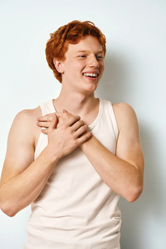 Young man with red hair and freckles smiling and holding chest in white tank top against white background in studio. Casual lifestyle portrait with happy male person.