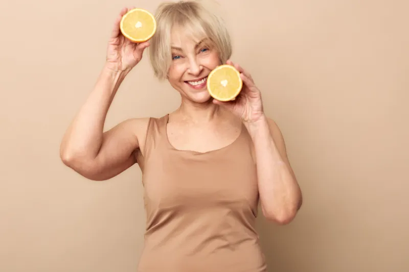 Happy mature woman in beige tank top holding two orange halves near face smiling against beige background. Concept of healthy lifestyle, natural beauty, and fresh fruit enjoyment.