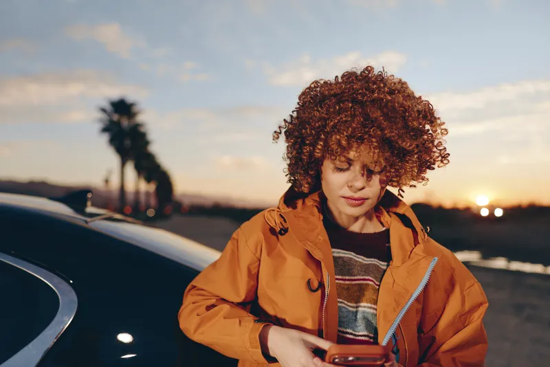 Woman lifestyle portrait near car at sunset, curly hair and orange jacket, looking at smartphone in hand, relaxed street atmosphere on evening road, warm golden light