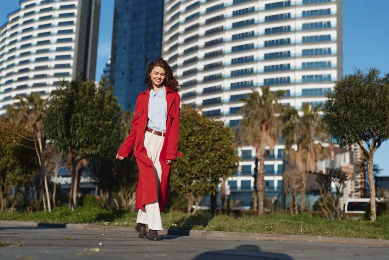 A stylish young woman with brown hair in a red coat walks along a sunny city street wearing white wide leg pants and a white blouse, with a confident smile.