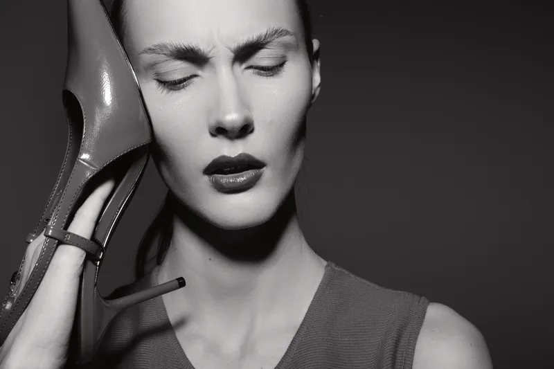 Close up of a stressed caucasian woman with pale skin holding a high heel shoe to her ear, eyes closed with a tense expression. Concept for frustration, fashion stress, and emotional discomfort.