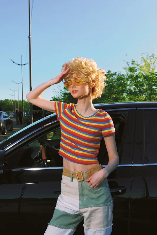 Woman outdoors in colorful striped top, retro style, sunlit moment with relaxed pose and wind in hair, vibrant background, casual confidence, warm natural tones