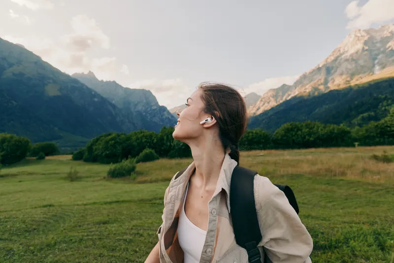Young woman with backpack gazes upward in a mountain valley, enjoying a sunny day amidst green meadows and distant peaks.