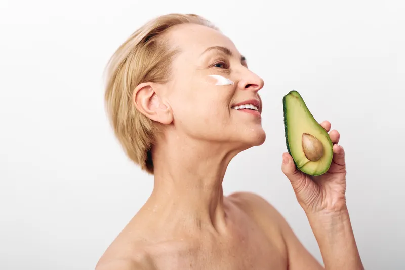 Mature woman with short blonde hair holds avocado near face with cream on cheek. Concept of natural skincare, healthy lifestyle, and anti aging beauty routine on white background.