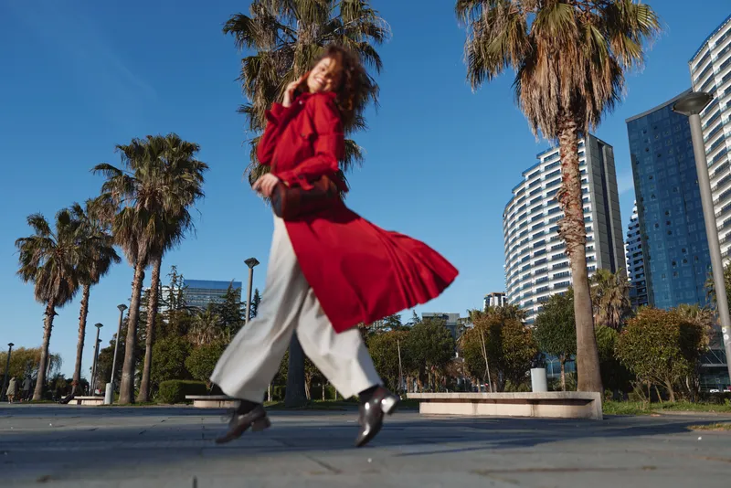 Young woman in a flowing red coat walks confidently along a sunny urban promenade lined with palm trees and modern buildings.