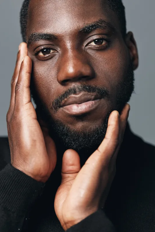 African man with dark skin and beard in creative conceptual visual style touching face with hands closeup portrait on gray background. Male model with thoughtful expression and textured skin.