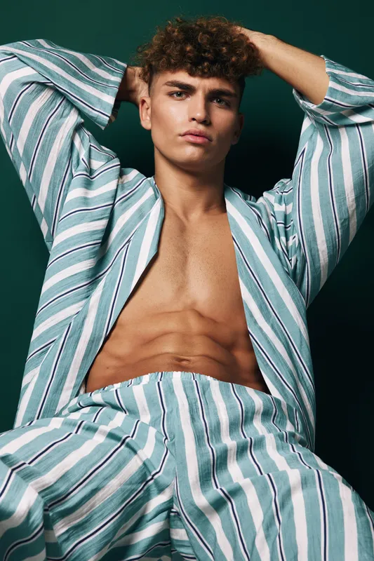 Man with curly hair and muscular body sitting with hands behind head wearing striped open shirt and pants against green background posing for camera indoors.