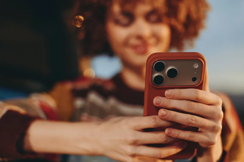 Woman lifestyle, holds smartphone for selfie, closeup hands and red phone, curly hair portrait, indoors on sofa, warm blurred background, soft focus bokeh
