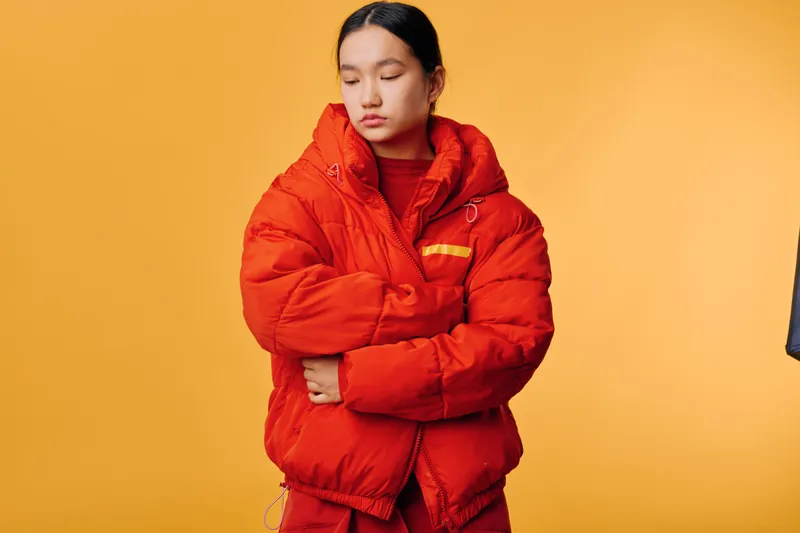 Young woman in red winter jacket and sweater standing with arms crossed against orange background. Female model posing with closed eyes wearing warm outerwear for cold weather.