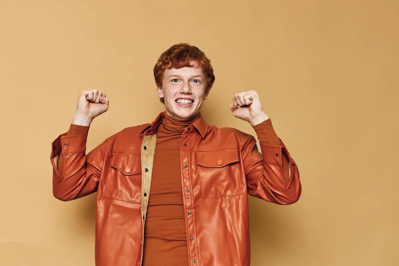 Redhead man with curly hair wearing orange jacket and turtleneck is smiling and raising fists in excitement on beige background for celebration or success.