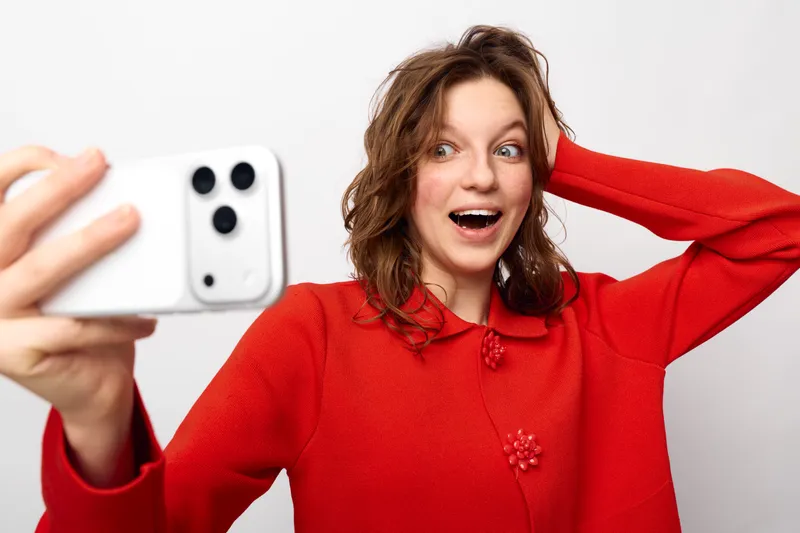 Young woman in red sweater takes a selfie with smartphone, smiling and surprised. White background, casual style, concept of social media, communication, and modern technology.