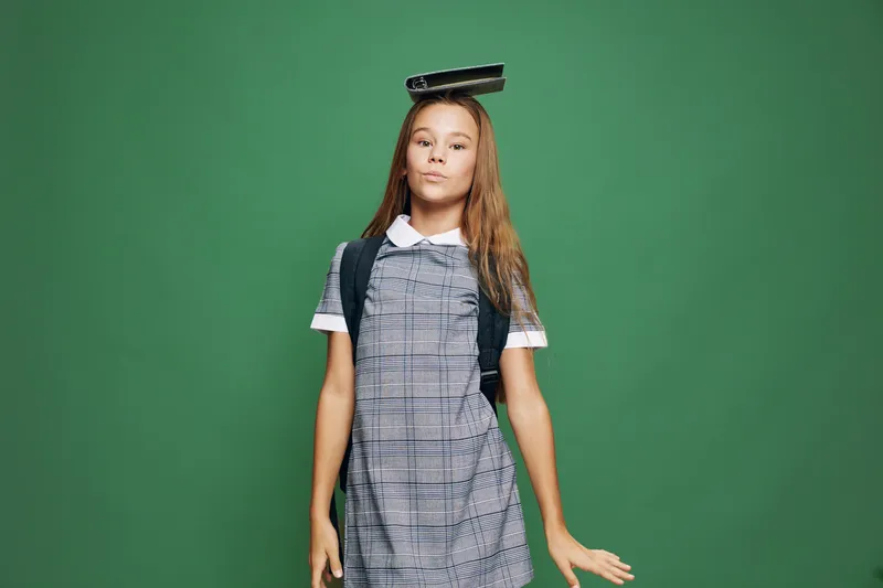 Schoolgirl with long hair and backpack wearing a checkered dress standing with notebook on head against green background. Child student posing confidently in studio.