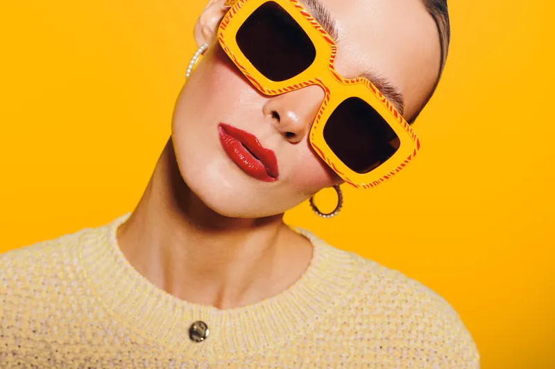 Close-up of a stylish woman wearing oversized orange sunglasses and red lipstick against a bright yellow background. Fashion concept with bold colors and modern accessories.