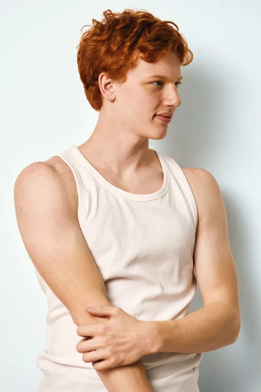 Young man with red hair wearing white sleeveless shirt stands sideways with arms crossed on white background. Casual style, natural light, and relaxed confident pose in studio.