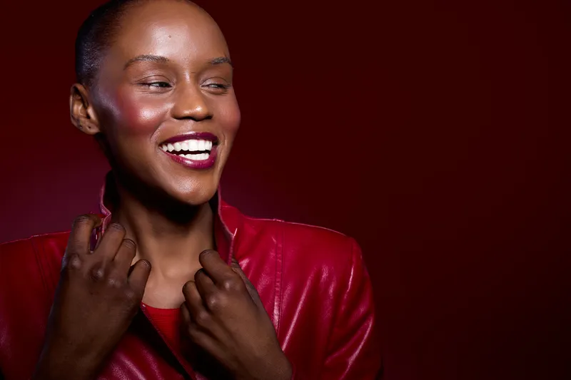 Smiling African American woman with dark skin wearing a red leather jacket looks sideways with joy. Studio portrait on red background. Concept of happiness, fashion, and beauty.
