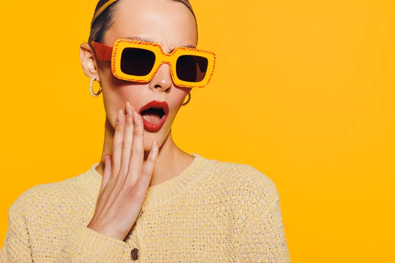 Surprised woman in yellow sweater and large orange sunglasses touches her cheek with open mouth on bright yellow background. Concept of shock, fashion, and colorful style.