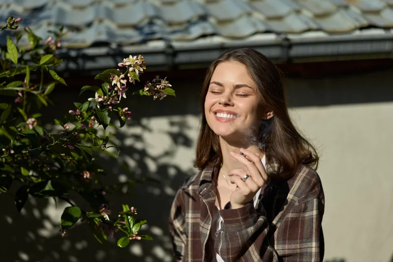 Bright woman lifestyle image shows a smiling figure outdoors near flowers and sunshine, wearing a plaid shirt, capturing a warm, relaxed moment in natural light