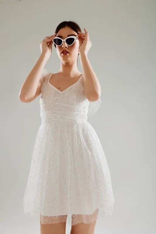 Young woman in a white dress adjusts white sunglasses. She poses confidently against a neutral background. Concept of beauty, fashion, and elegance in modern style.